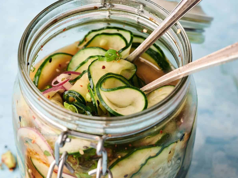 A glass jar filled with sliced cucumbers, red onions, and herbs in pickling liquid, with two metal forks placed inside.