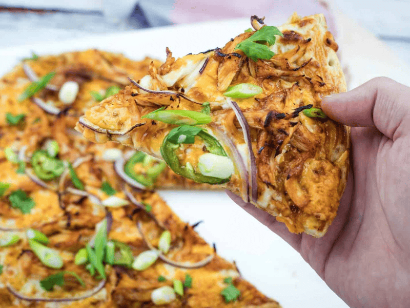 A hand holds a slice of pizza topped with shredded chicken, jalapeño slices, red onions, and green onions; the rest of the pizza is visible on a white surface.