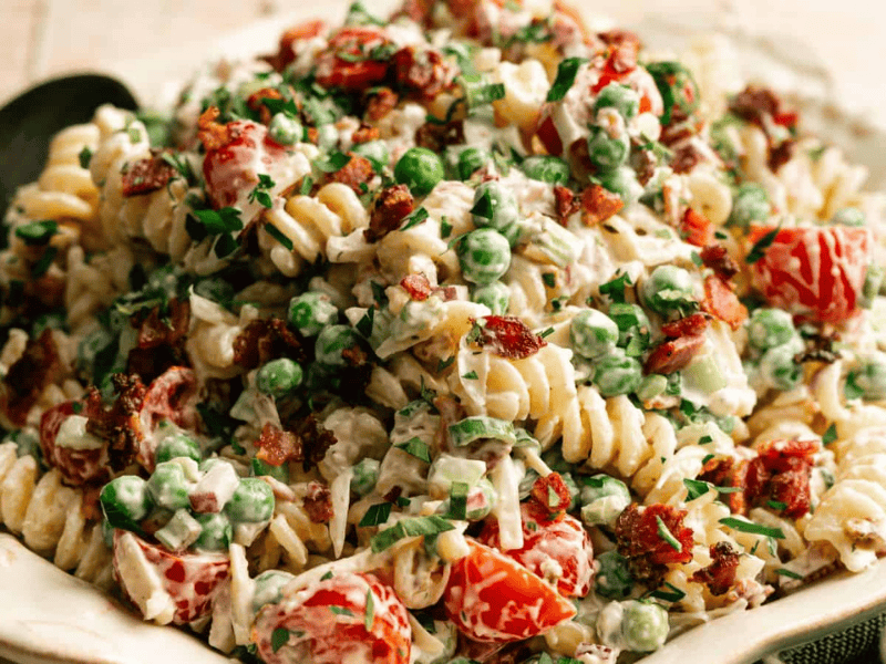 A bowl of rotini pasta salad mixed with peas, cherry tomatoes, bacon bits, chopped herbs, and a creamy dressing, with a black serving spoon beside the dish.