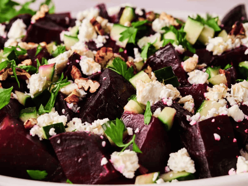 A close-up of a salad featuring chopped beets, cucumber, feta cheese, walnuts, and fresh herbs on a white plate.
