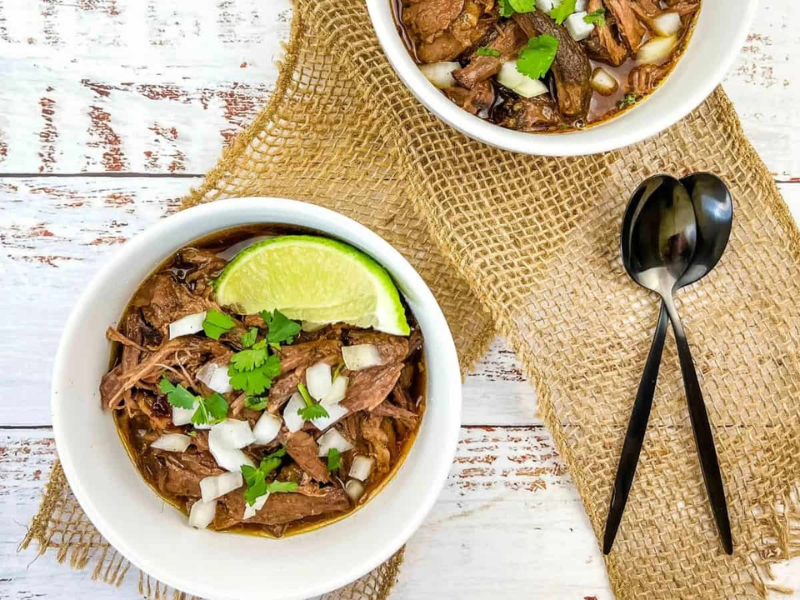 Two white bowls filled with slow cooker birria garnished with fresh lime wedges.
