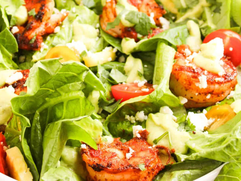 A bowl of mixed salad with grilled shrimp, avocado, cherry tomatoes, lettuce, feta cheese, and a creamy dressing.