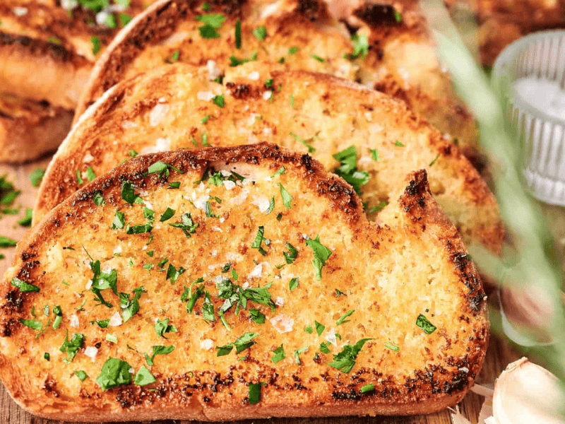 Slices of garlic bread topped with chopped parsley and sea salt sit enticingly on a wooden board, accompanied by a small bowl of salt and fresh cloves of garlic.