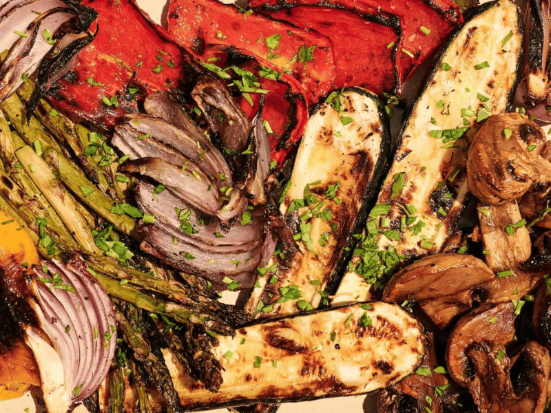 A platter of grilled vegetables including zucchini, mushrooms, asparagus, bell peppers, and red onions, garnished with chopped herbs.