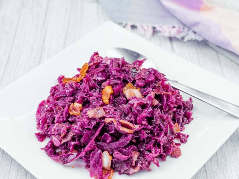A serving of cooked red cabbage with pieces of bacon and carrots on a white square plate, with a spoon beside the food.