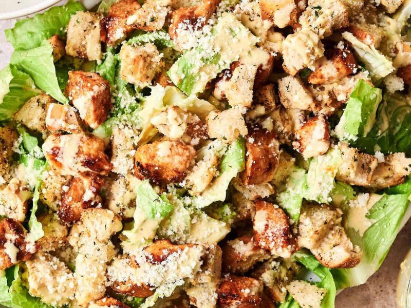 A Caesar salad topped with grilled chicken pieces, croutons, and grated cheese on a bed of romaine lettuce. Dressing is served on the side.