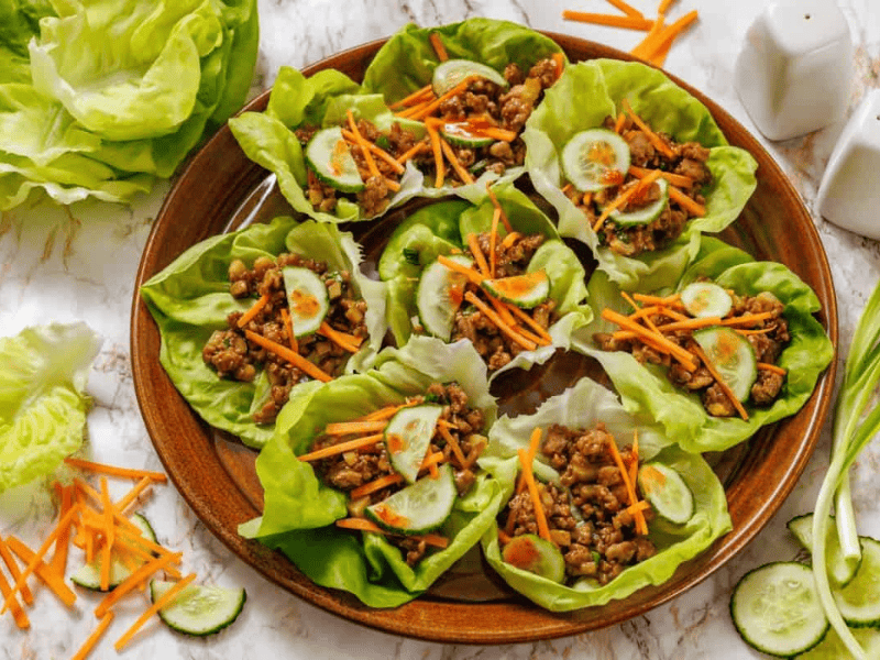 A wooden plate with Chicken lettuce wraps surrounded by extra lettuce leaves and vegetables on a marble surface.