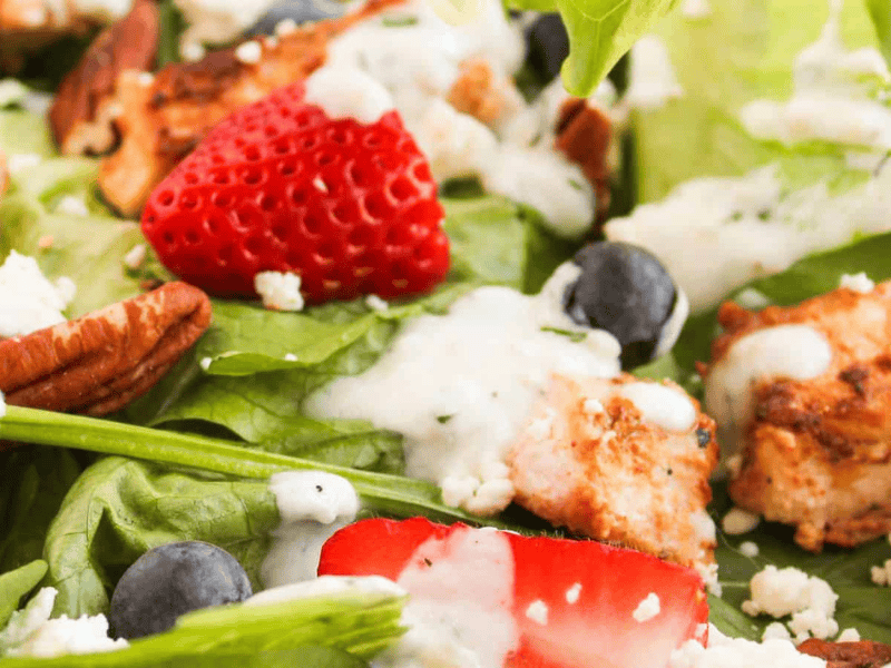 Close-up of a salad with grilled chicken, strawberries, blueberries, pecans, feta cheese, and a creamy dressing on a bed of leafy greens.