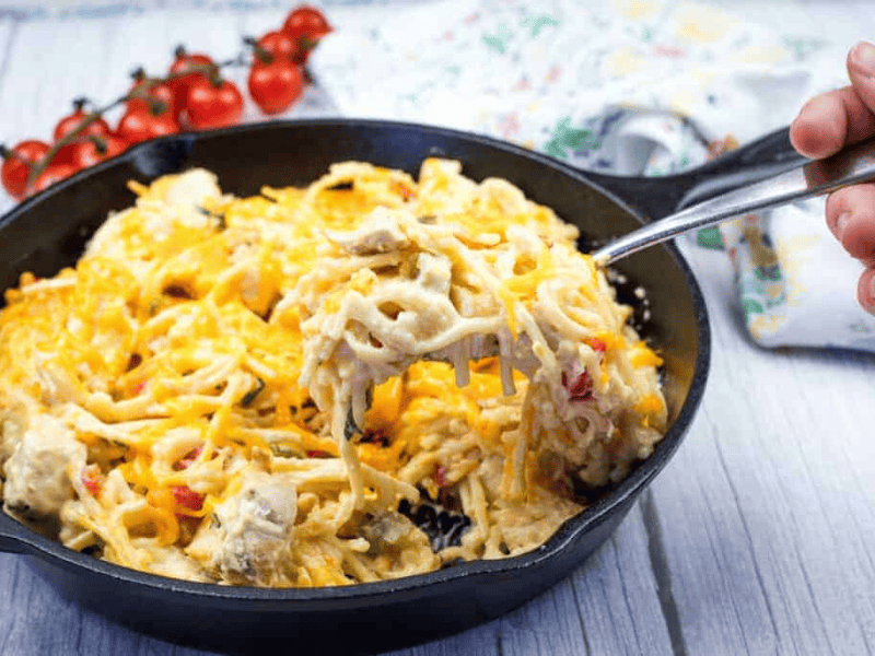 A hand holding a fork with a serving of chicken spaghetti from a cast-iron skillet.