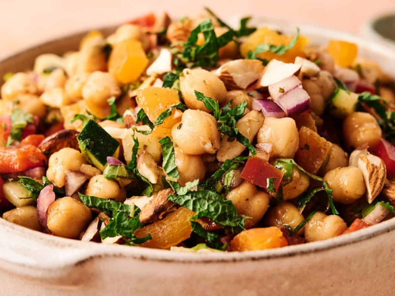 A ceramic bowl filled with chickpea salad, including chopped red onion, yellow peppers, tomatoes, almonds, and fresh herbs.