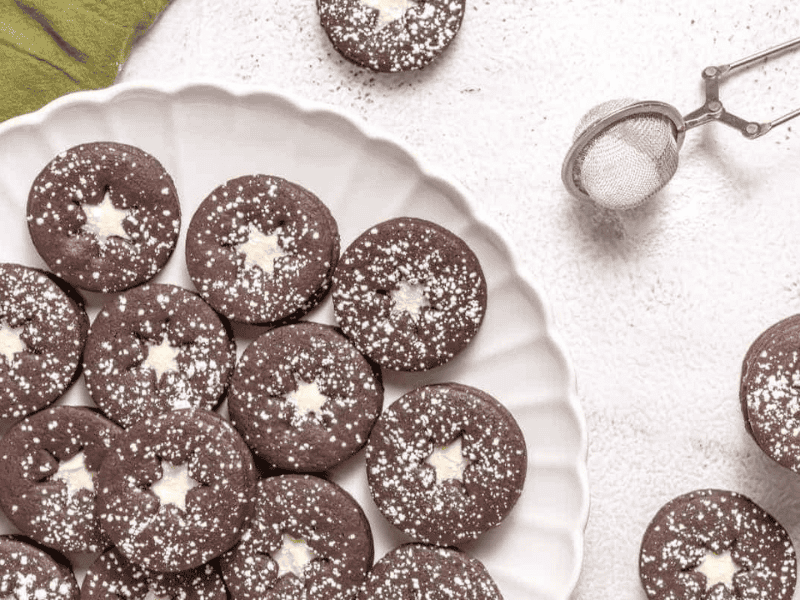 A plate of round chocolate cookies with star-shaped centers, dusted with powdered sugar. A metal sifter with powdered sugar is on the side.