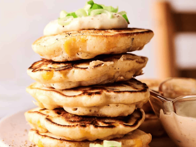 A stack of corn fritters topped with a dollop of cream and chopped green onions, served on a plate with a bowl of dipping sauce nearby.