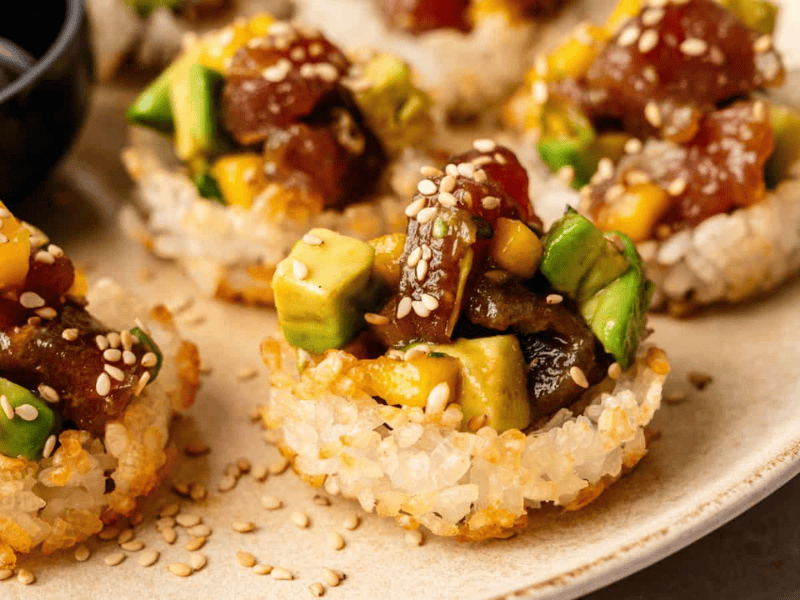 Close-up of crispy rice rounds topped with diced tuna, avocado, mango, and sesame seeds, served on a beige plate with a small dish of soy sauce.