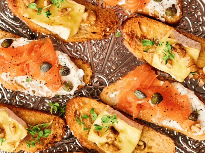 Toasted bread topped with smoked salmon, capers, cheese, walnuts, and herbs on a decorative silver tray.