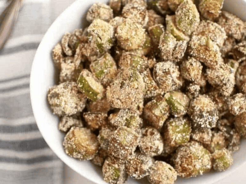 A white bowl filled with crispy, breaded and fried okra pieces, placed on a light surface next to a striped cloth napkin.
