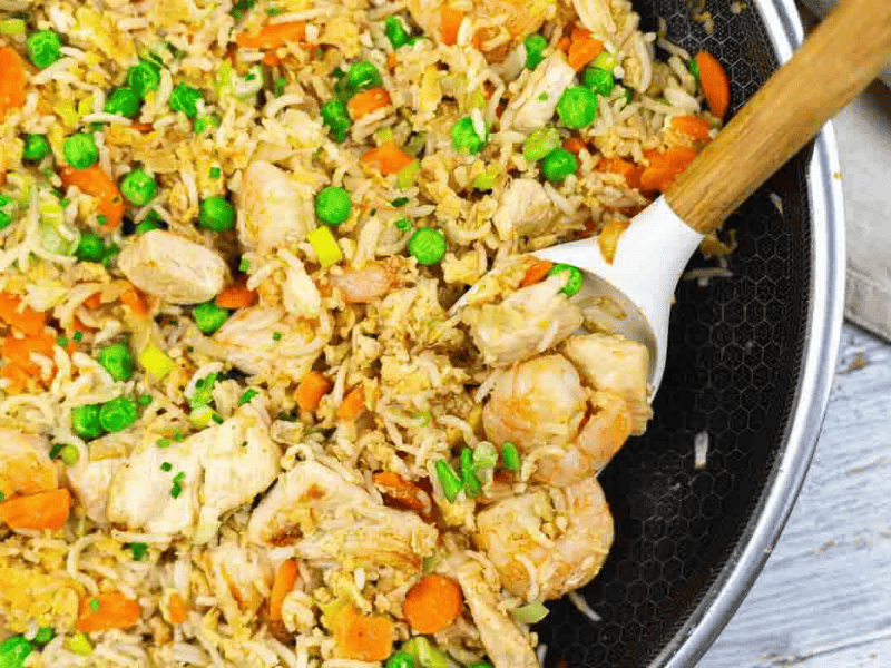 A pan filled with fried rice, shrimp, chicken pieces, carrots, peas, and scrambled eggs, with a white spoon for serving.