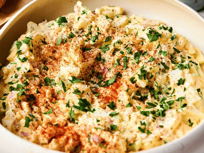 A bowl of creamy egg salad topped with chopped parsley and paprika, set on a light-colored surface.