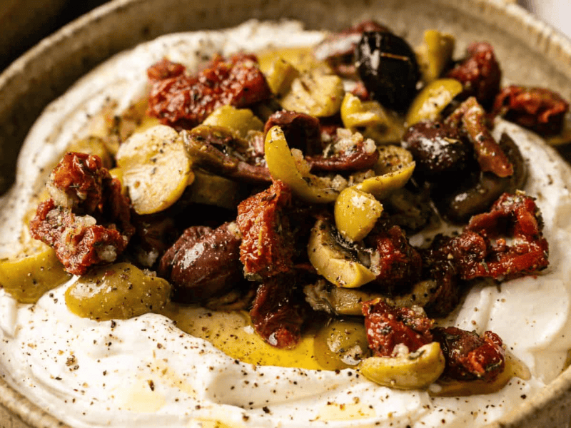 A bowl of whipped feta cheese topped with mixed olives, sun-dried tomatoes, herbs, and olive oil.