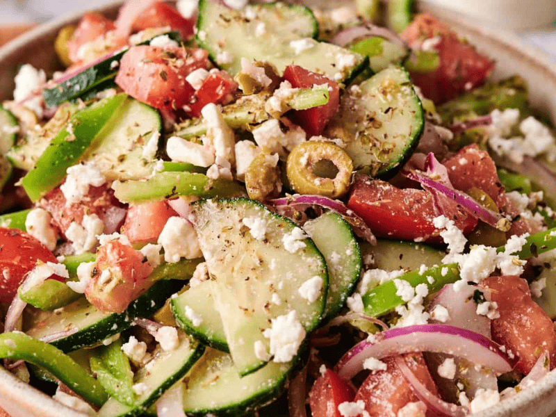 A bowl of Greek salad with sliced cucumbers, tomatoes, red onions, green bell peppers, olives, and crumbled feta cheese.