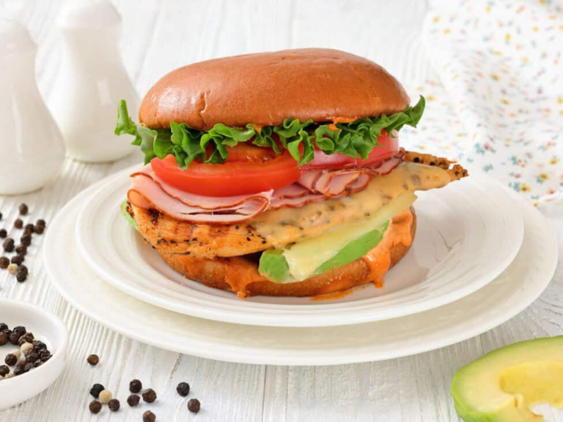 A Grilled California Chicken Burger served on a white plate with peppercorns and a napkin nearby.