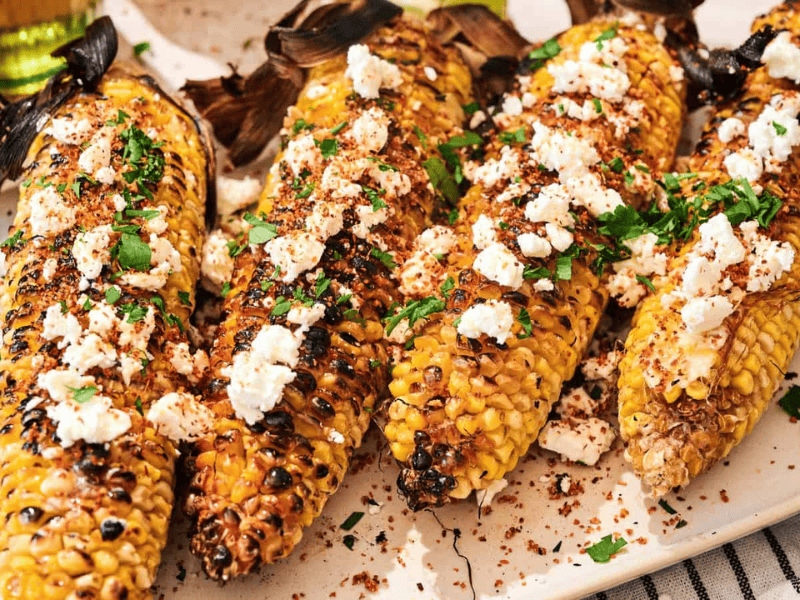Four grilled corn cobs topped with crumbled cheese, chopped herbs, and spices are arranged on a white plate.
