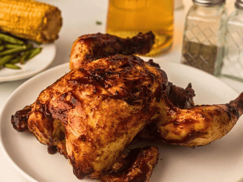 A roasted chicken with barbecue sauce on a white plate, with green beans, corn on the cob, a glass of beer, and salt and pepper shakers in the background.