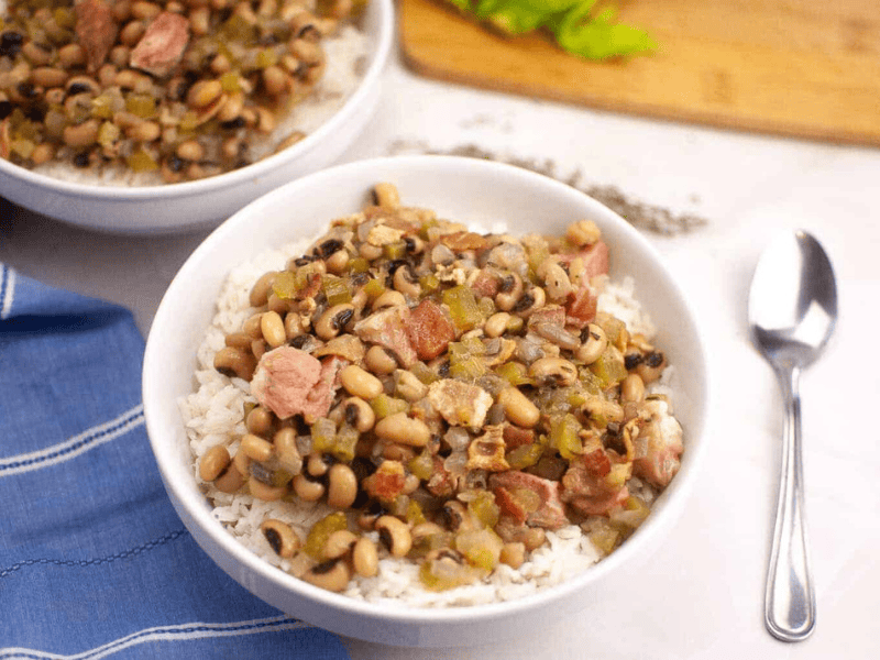 A bowl of Hoppin&rsquo; John served over white rice, with a spoon and a blue napkin nearby.
