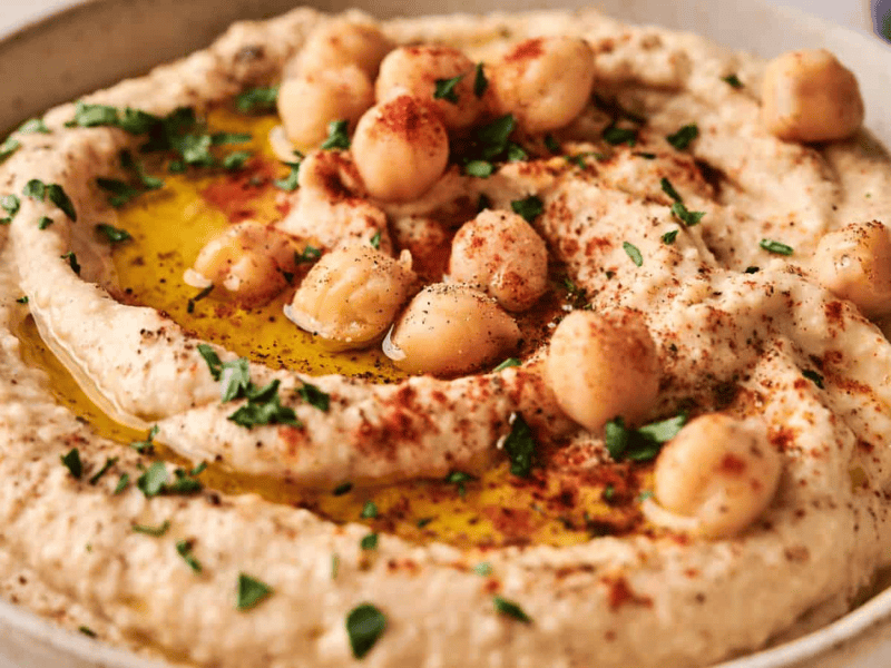 A bowl of hummus garnished with whole chickpeas, chopped parsley, paprika, and a drizzle of olive oil.