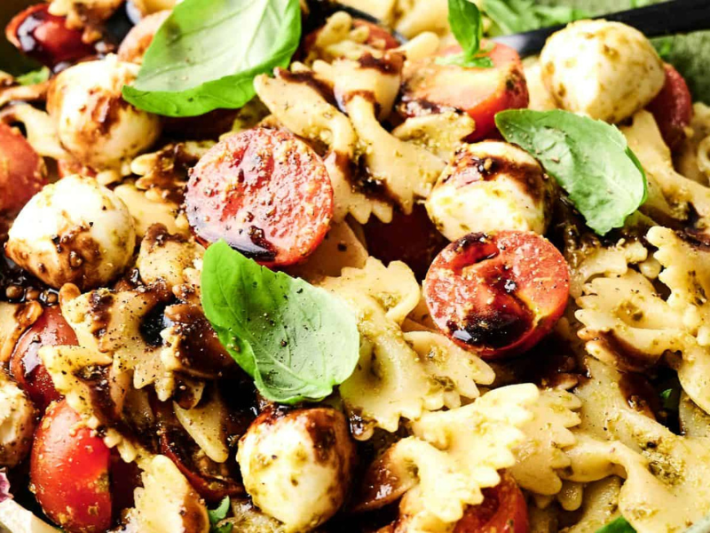 Bowtie pasta salad with cherry tomatoes, mozzarella balls, fresh basil leaves, and a drizzle of balsamic glaze in a green bowl.