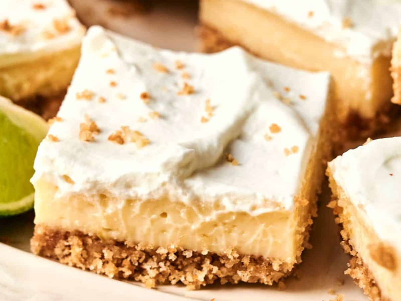 A close-up of dessert bars with a crumbly crust, creamy filling, and whipped topping, garnished with lime slices in the background.