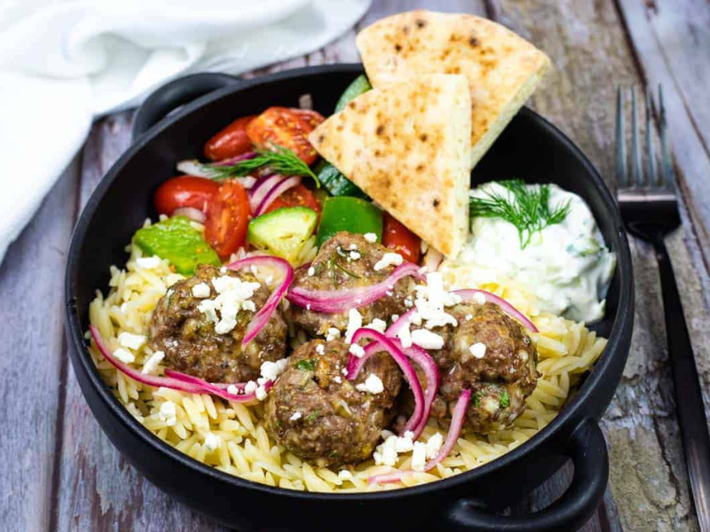 A black bowl with orzo pasta, meatballs topped with feta and pickled onions, Greek salad, pita bread, and a side of tzatziki sauce on a rustic wooden table.