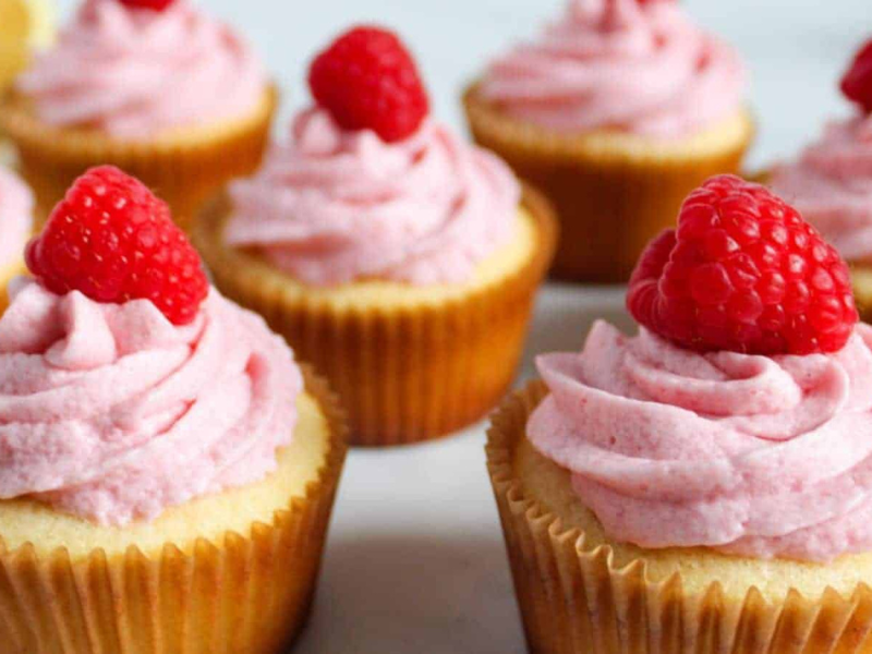 Vanilla and lemon cupcakes with swirled pink raspberry buttercream frosting, each topped with a fresh raspberry, arranged closely together.