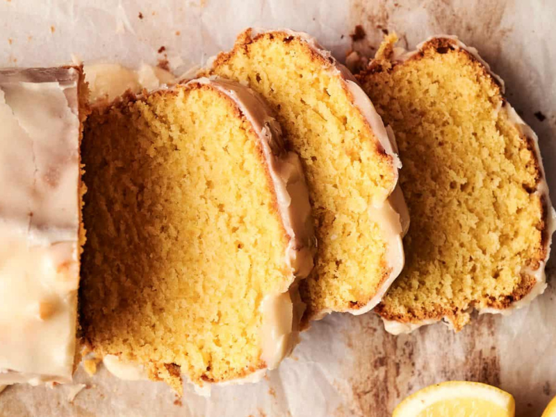 Sliced lemon cake with glaze on parchment paper, next to lemon slices.