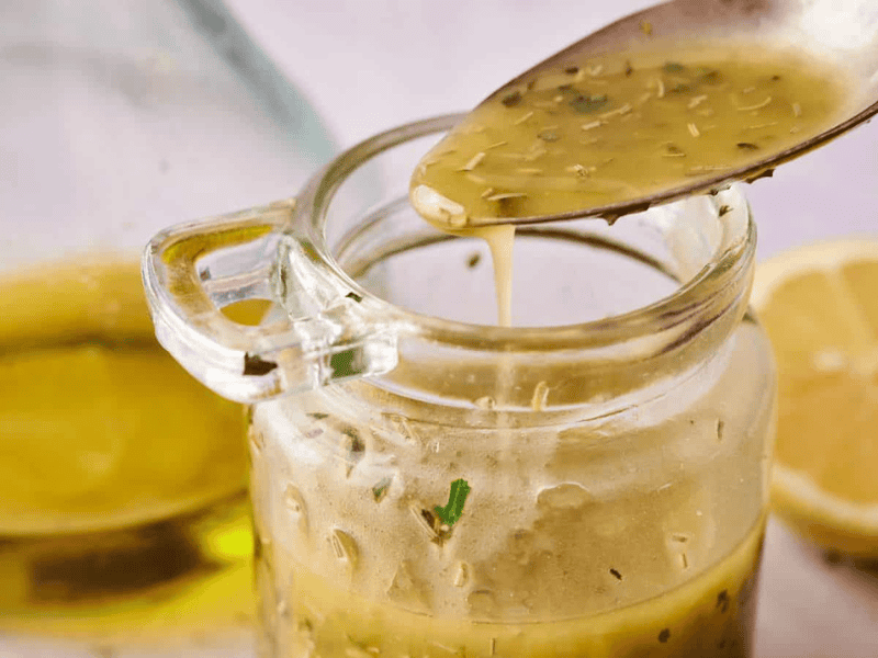 A spoon drizzles vinaigrette from a glass jar, with herbs visible in the dressing; a halved lemon and a bottle of oil are in the background.