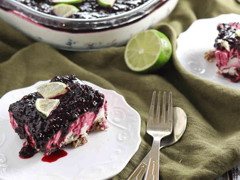 A slice of berry-topped dessert with lime wedges sits on a white plate next to a fork and knife, with more dessert and a halved lime in the background.