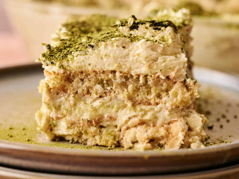 A close-up of a slice of tiramisu topped with green matcha powder, served on a ceramic plate.