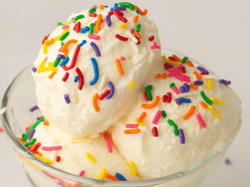 A glass bowl with two scoops of vanilla ice cream topped with colorful rainbow sprinkles, set against a plain white background.