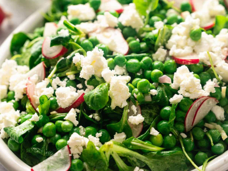 A close-up of a salad with green peas, leafy greens, sliced radishes, and crumbled feta cheese on a white plate.