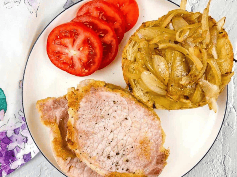 Peameal bacon on a plate with cut tomatoes and cooked onions.