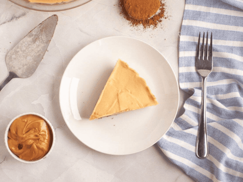 A slice of Peanut Butter & Chocolate Pie on a white plate, with a fork, a small bowl of peanut butter, a baking tool, and a striped napkin nearby.