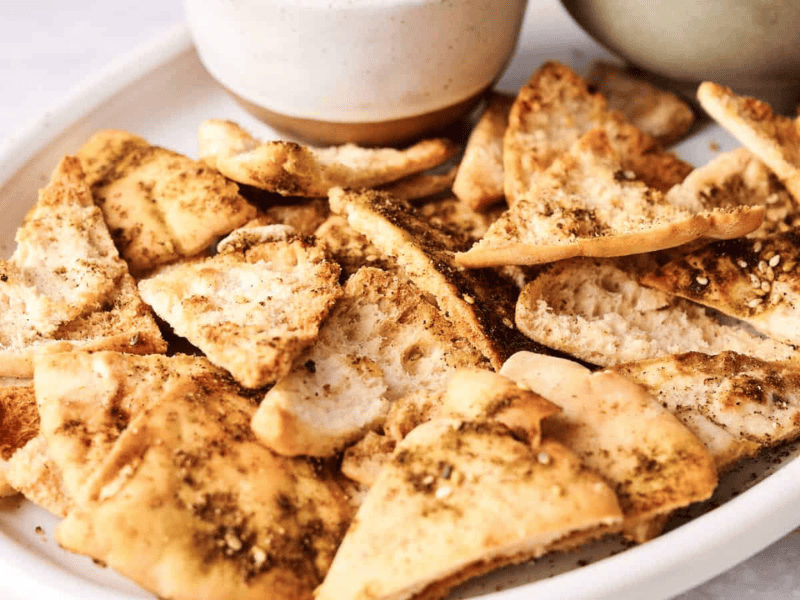 A white oval plate with crispy pita chips seasoned with spices, placed next to ceramic bowls.