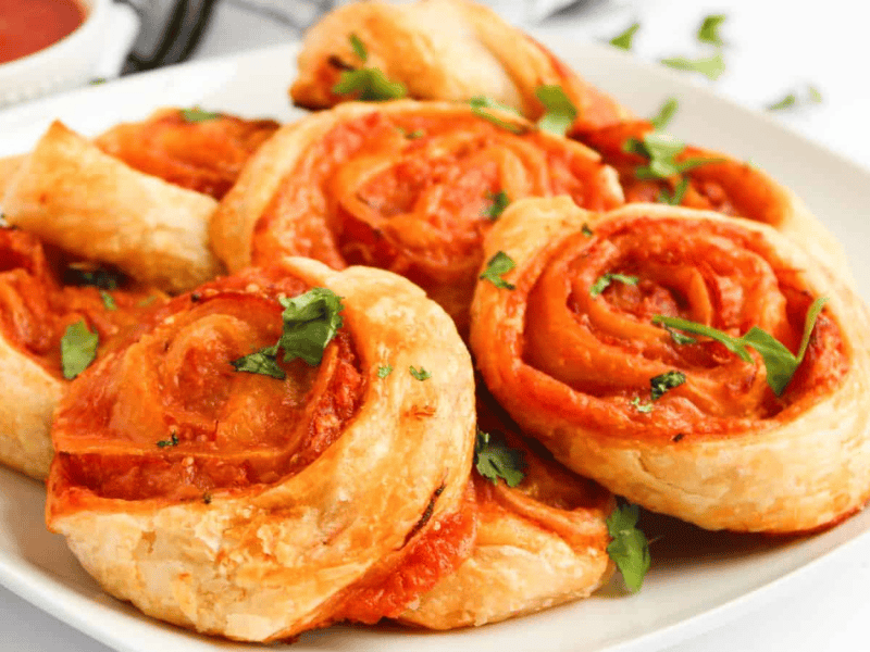 A plate of golden-brown pinwheel pastries filled with tomato sauce and garnished with fresh herbs. A small bowl of sauce is in the background.