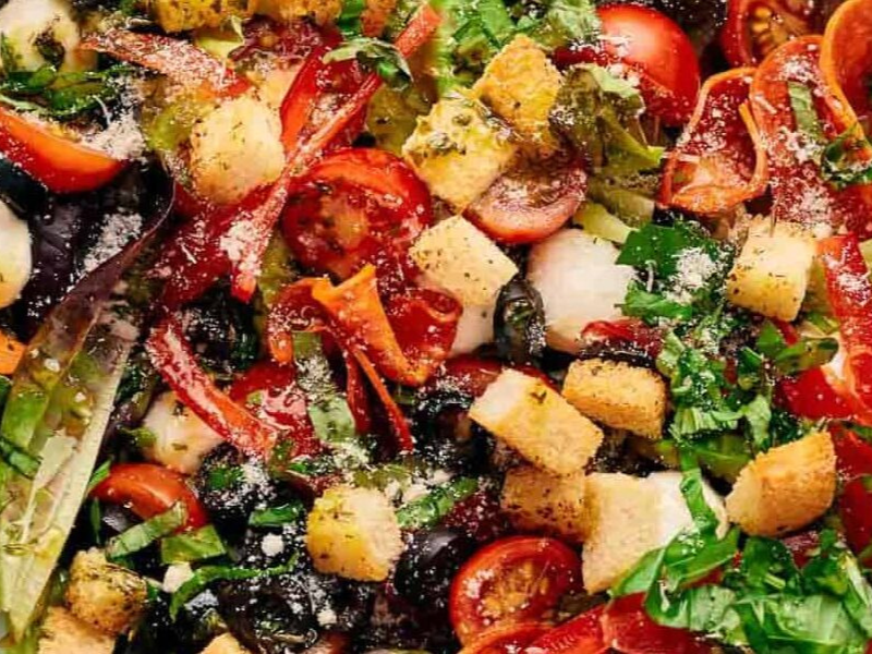 A close-up of a fresh salad with cherry tomatoes, croutons, lettuce, olives, sliced peppers, greens, and grated cheese.