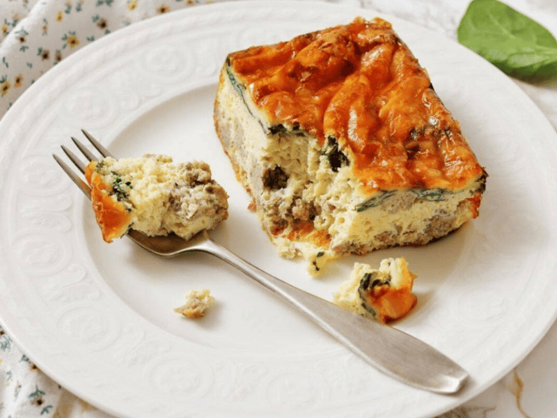 A slice of Sausage, Spinach, and egg casserole on a white plate, with a fork holding a bite-sized piece.