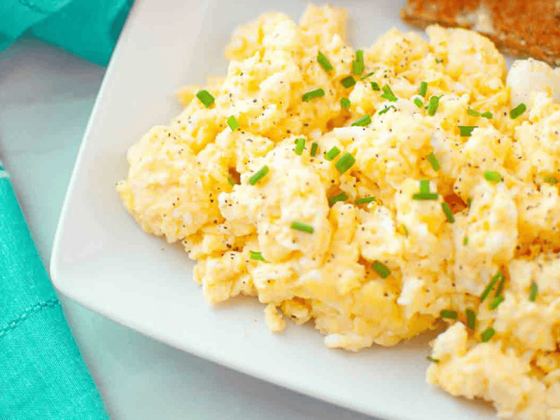 Scrambled eggs garnished with chopped chives and black pepper, served on a white plate next to a piece of toast.