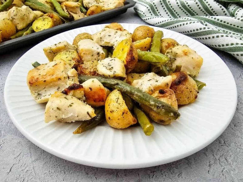 A white plate with roasted chicken pieces, halved baby potatoes, and green beans, with a baking tray and striped towel in the background.
