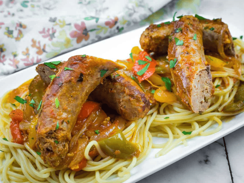 Slow Cooker Sausage & Peppers served over spaghetti garnished with herbs, on a white rectangular plate.