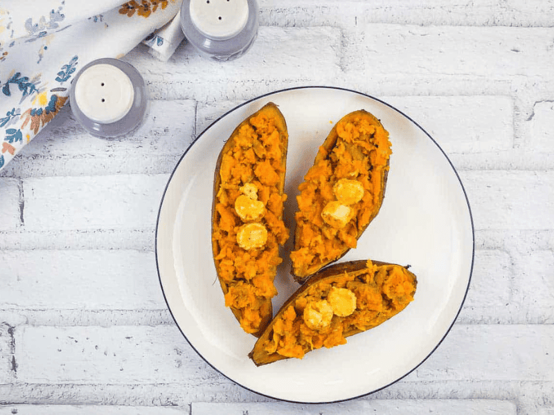 Three halves of roasted sweet potatoes topped with butter on a white plate, set on a white brick surface with a floral napkin and salt and pepper shakers nearby.