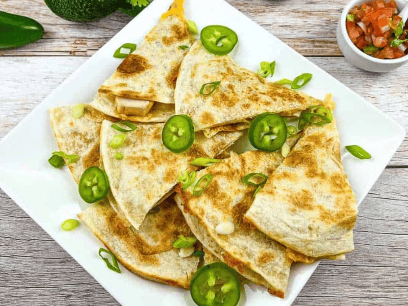 A white plate with sliced quesadillas topped with sliced green jalapeños and chopped green onions. A small bowl of salsa sits nearby on a wooden table.