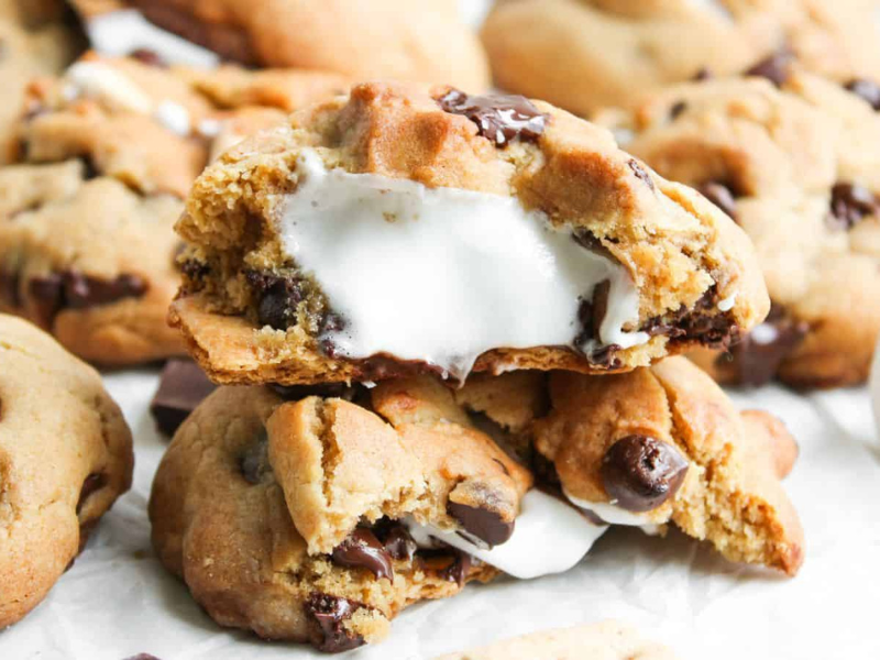 A close-up of chocolate chip cookies filled with melted marshmallow, with one cookie broken open to show the gooey center.